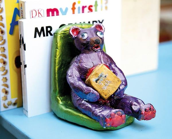 Sevimli ayı kitap desteği: yeşil koltukta kitap tutan mor metalik renkli kil ayı, kitapların yanında