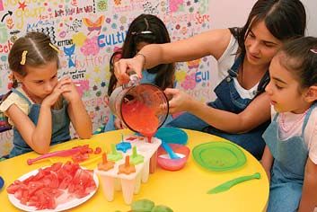 Kırmızı meyve karışımı sürahiden plastik loli buz kalıplarına dökülüyor, masada karpuz dilimleri ve izleyen çocuklar