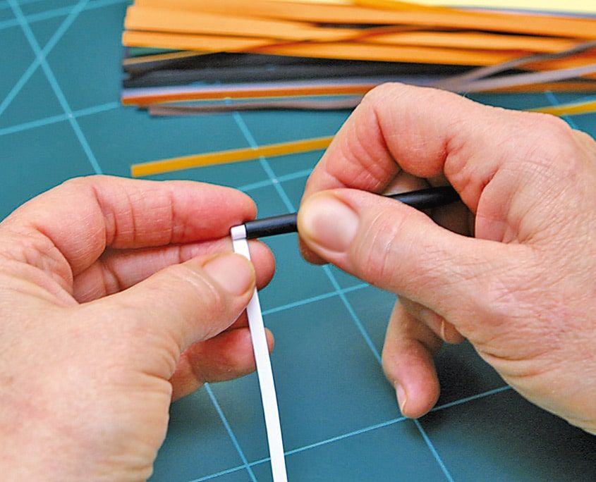 Beyaz quilling şeridi yarıklı çubuğun ucuna takılıyor, arkada renkli kağıt şeritler