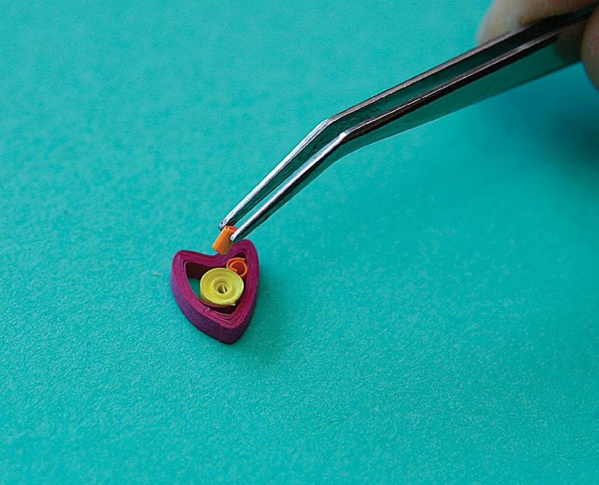 Cımbızla tutulan kalp biçimli bordo quilling parçasının içine turuncu küçük rulo ekleniyor