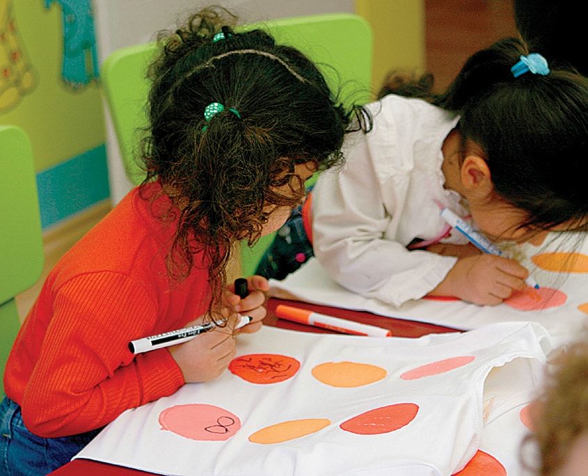 Masaya serilmiş tişörtlerdeki turuncu yuvarlakların içine kumaş kalemiyle yüz çizen çocuklar
