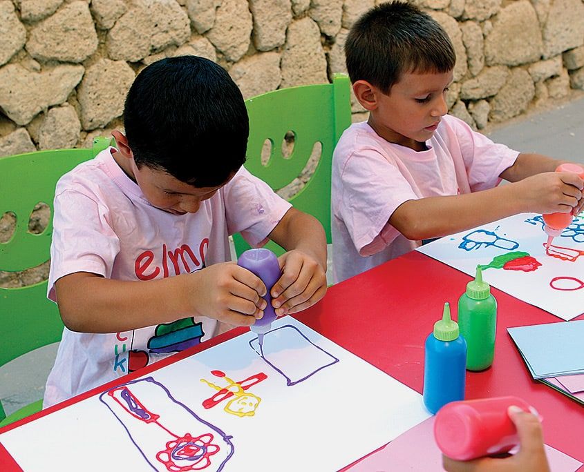 Masada çalışan çocuklar sıkma şişelerdeki renkli kabaran boyayla kağıda şekiller çiziyor