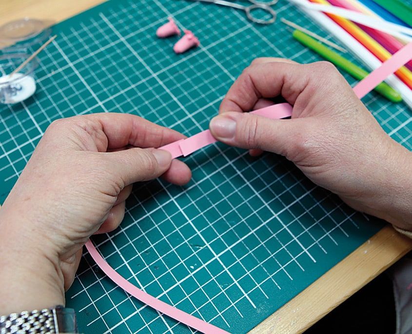 Bir cm genişliğindeki pembe quilling şeridi iki elle gerilerek düzleştiriliyor, masada kesim altlığı