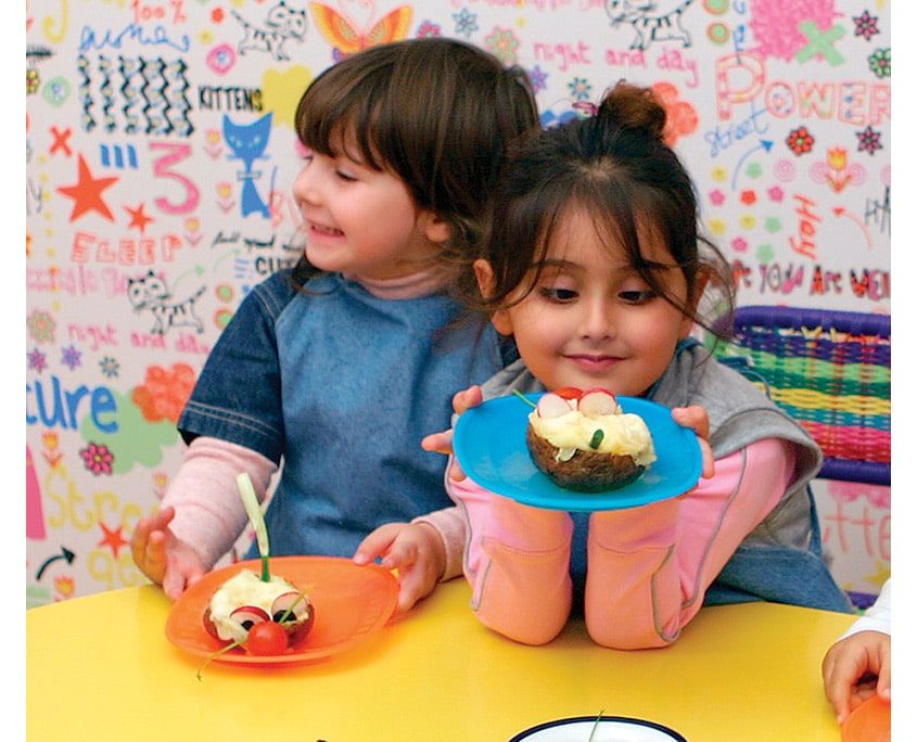 Önlüklü iki çocuk mavi ve turuncu tabaklardaki hazırladıkları fare kumpirleri gösteriyor