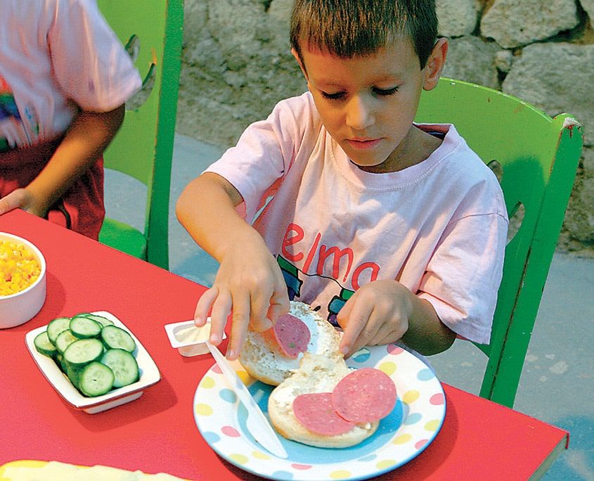 Krem peynir sürülmüş ekmeğe salam dilimleri yerleştiriliyor, yanda kasede dilimlenmiş salatalık