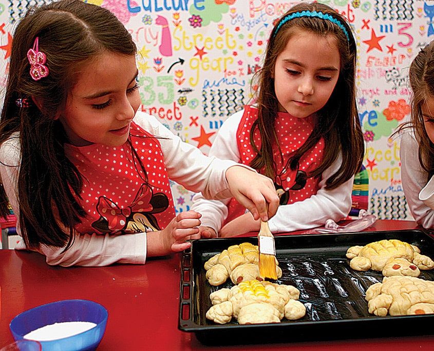 Önlüklü çocuklar tepsideki kaplumbağa ekmeklerin üzerine fırçayla yumurta sarısı sürüyor
