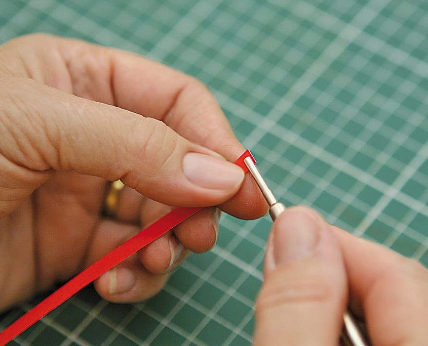 Kırmızı quilling şeridinin ucunu dolama aletinin yarığına geçiren parmaklar