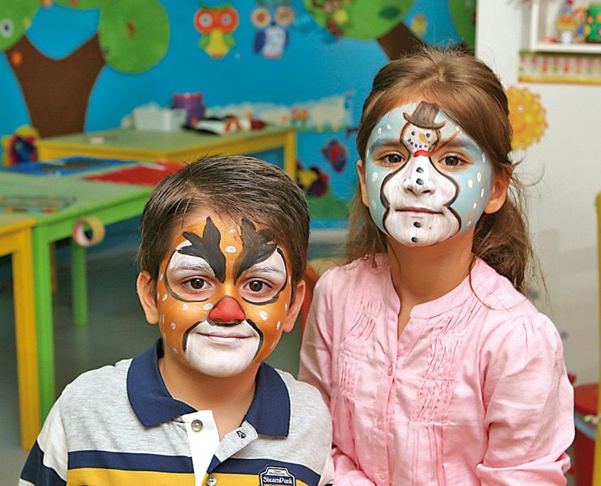 Geyik yüz boyamalı bir çocuk ile maymun yüzü boyanmış bir çocuk atölye sınıfında yan yana