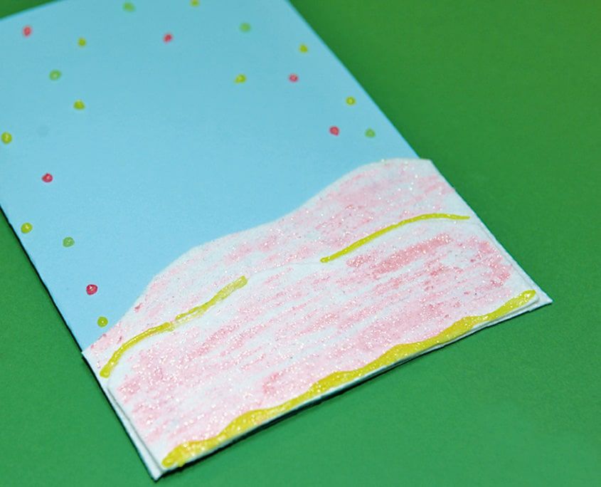 Mavi kartın alt kısmına pembe ve simli pastelle boyanmış kar tepeleri, üstte renkli kar noktaları