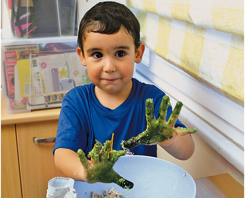 Kasenin başındaki çocuk yeşil hamura bulanmış iki elini havaya kaldırıp gösteriyor