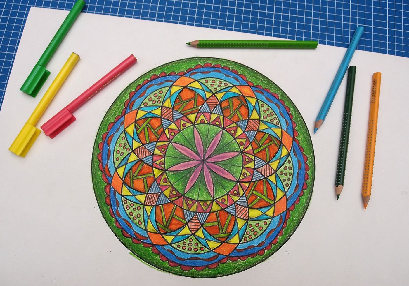 Yeşil, turuncu, mavi ve pembe tonlarda boyanmış yıldız merkezli mandala, çevresinde kalemler