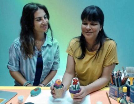 Taş boyama canlı atölyesinden kare: iki eğitmen masada, saksılara yerleştirilmiş boyalı taşlar