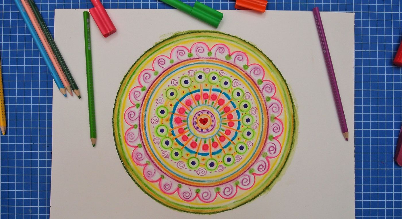 Kağıda çizilmiş halkalı mandala renkli kalemlerle boyanıyor: pembe spiraller, yeşil ve mavi noktalar