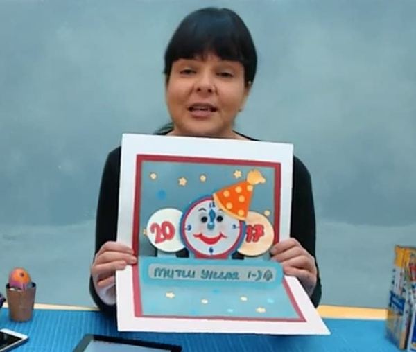 Yeni yıl kartları canlı atölyesinden kare: eğitmen parti şapkalı gülen saat figürlü açılır kartı gösteriyor