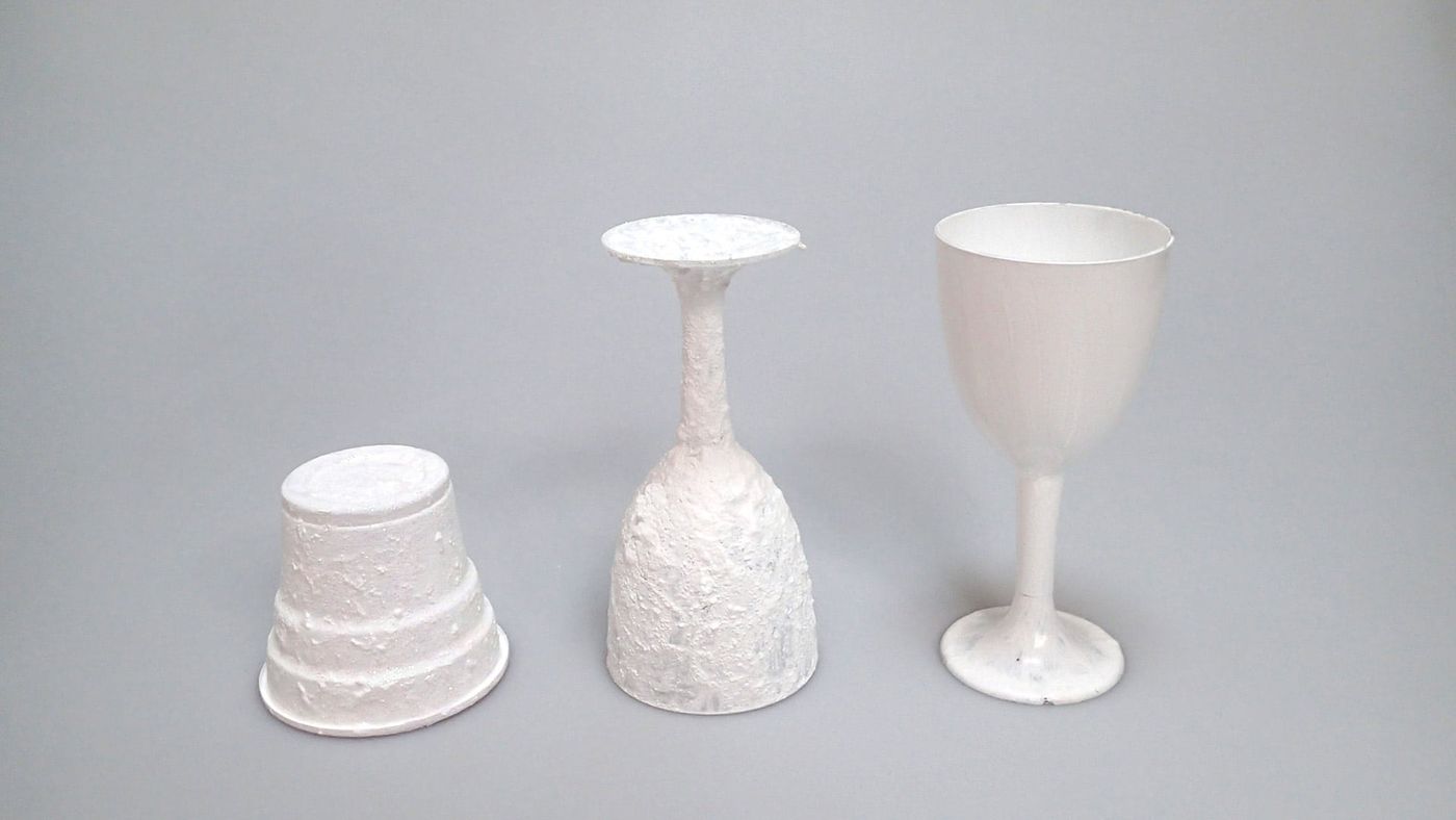 Kabartma dokulu beyaz boyayla kaplanmış bardak ve ters kadeh, yanında boyanmamış plastik kadeh