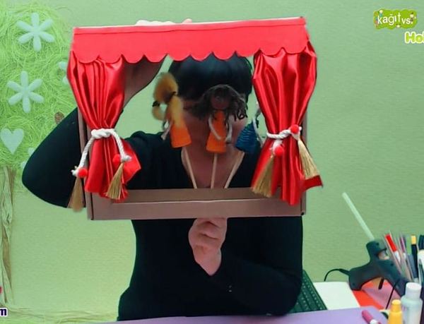 Canlı atölye yayınından kare: eğitmen kırmızı perdeli karton kutu sahnenin arkasından çubuk kuklaları oynatıyor