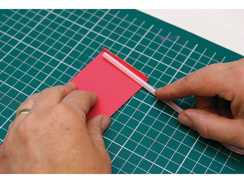 Kesim altlığında pembe kağıt şeridin kenarına plastik pipet yerleştirilip sarılmaya başlanıyor