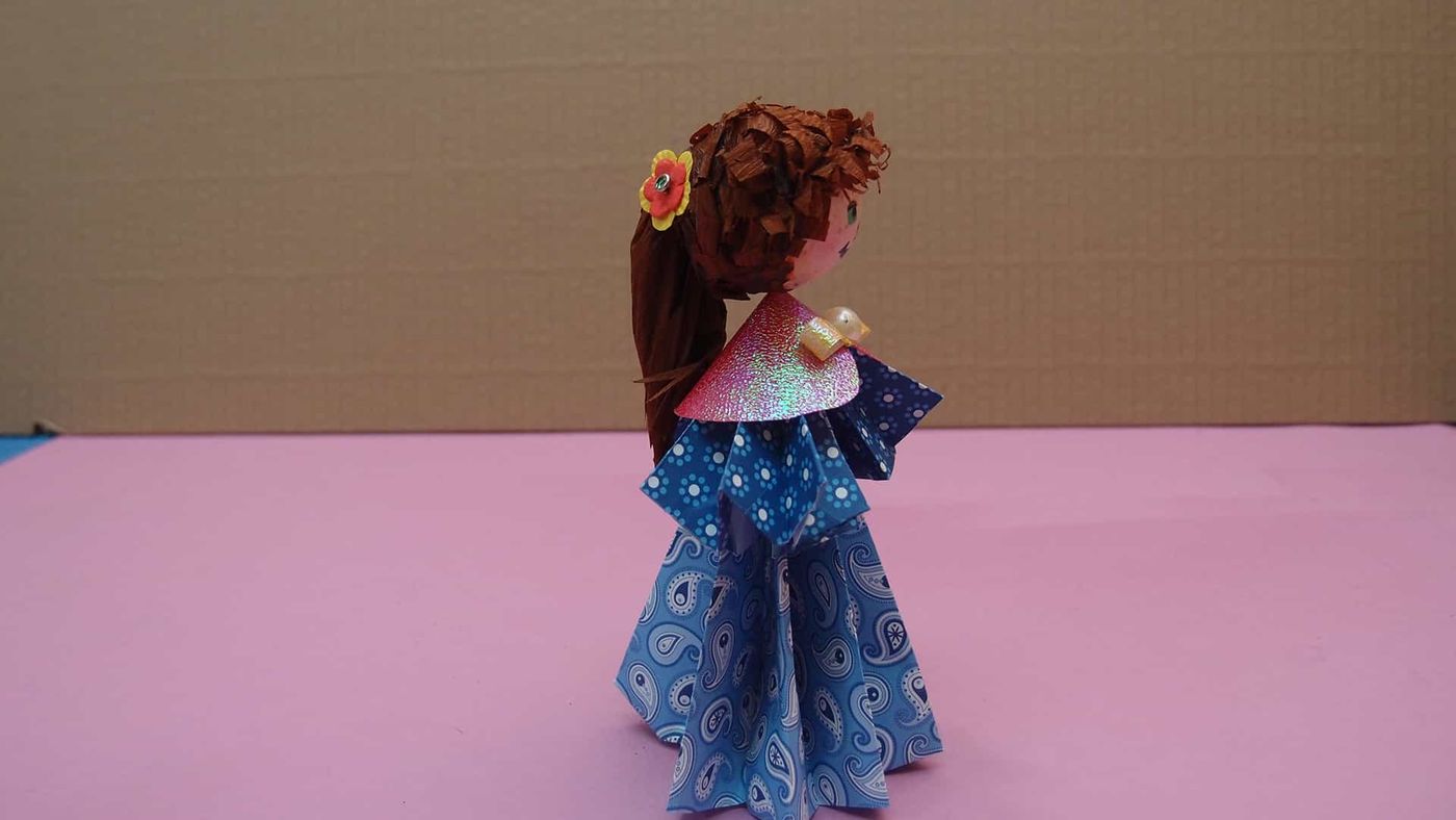 Kıvırcık saçlı origami bebek yandan: saçında çiçek, arkaya uzanan uzun kahverengi saç ve mavi elbise