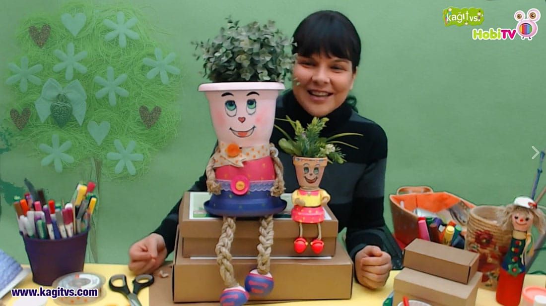 Neşeli saksı: canlı atölye yayınından kare, eğitmen yüzü boyalı, bitkili iki saksı bebeği gösteriyor
