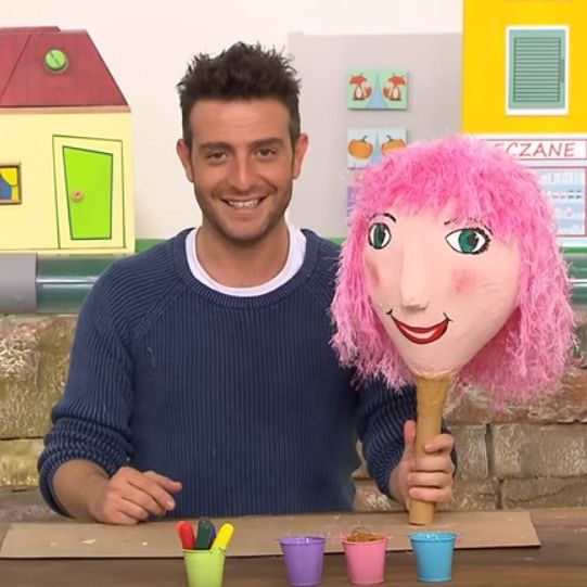 Balon Kafalar: televizyon stüdyosunda sunucu, pembe tüylü saçlı kağıt hamuru balon kafa kuklayı gösteriyor