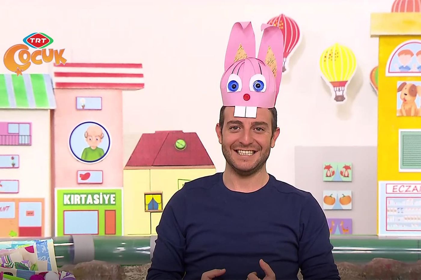 Stüdyoda sunucu başında pembe tavşan şapkayla konuşuyor, arkada renkli ev ve balon dekorları