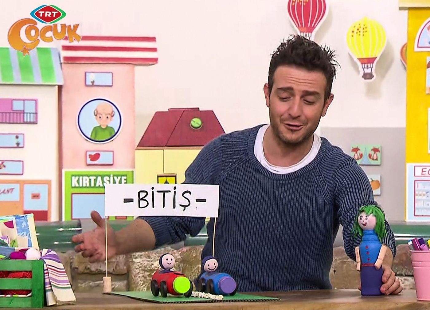Program karesi: sunucu masadaki Bitiş tabelalı pisti, şişe arabaları ve şişe bebeği gösteriyor