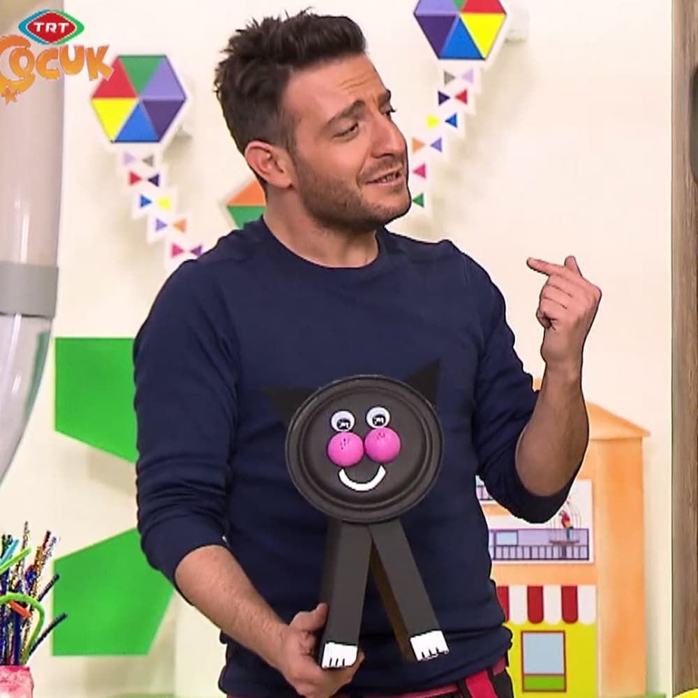 Sunucu siyah kedi dosyalığı tutarken diğer eliyle onu gösteriyor, arkada renkli dekor