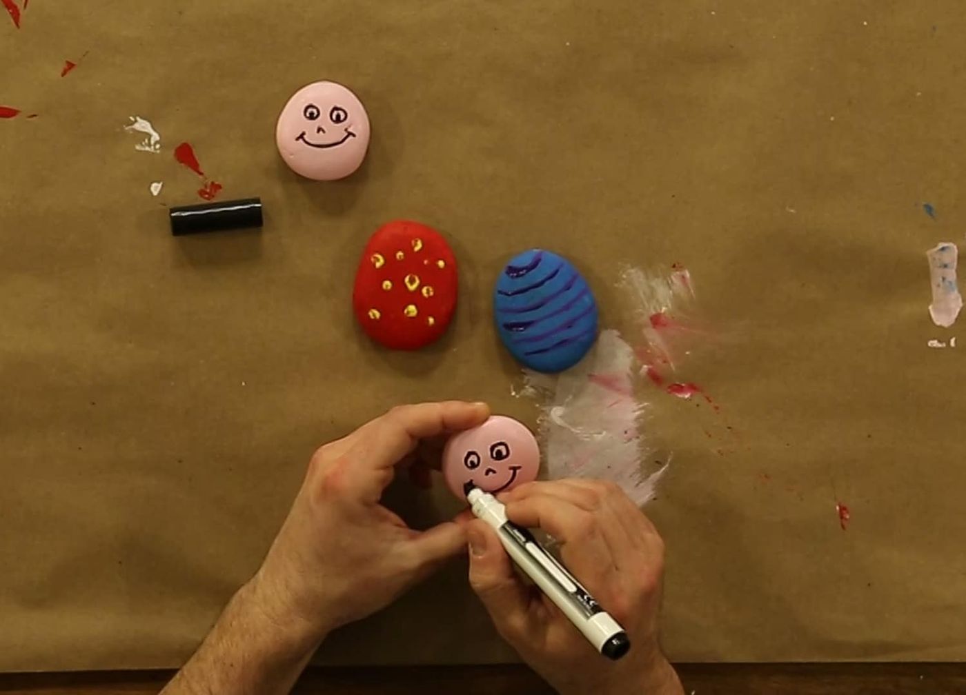Pembe boyanmış taşa keçeli kalemle yüz çiziliyor, yanda kırmızı ve mavi desenli gövde taşları