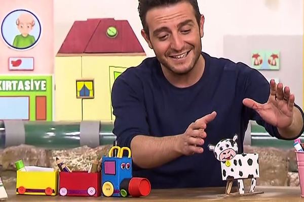 Programdan kare: sunucu masadaki renkli kutu tren kalemliği ve karton inek figürünü gösteriyor