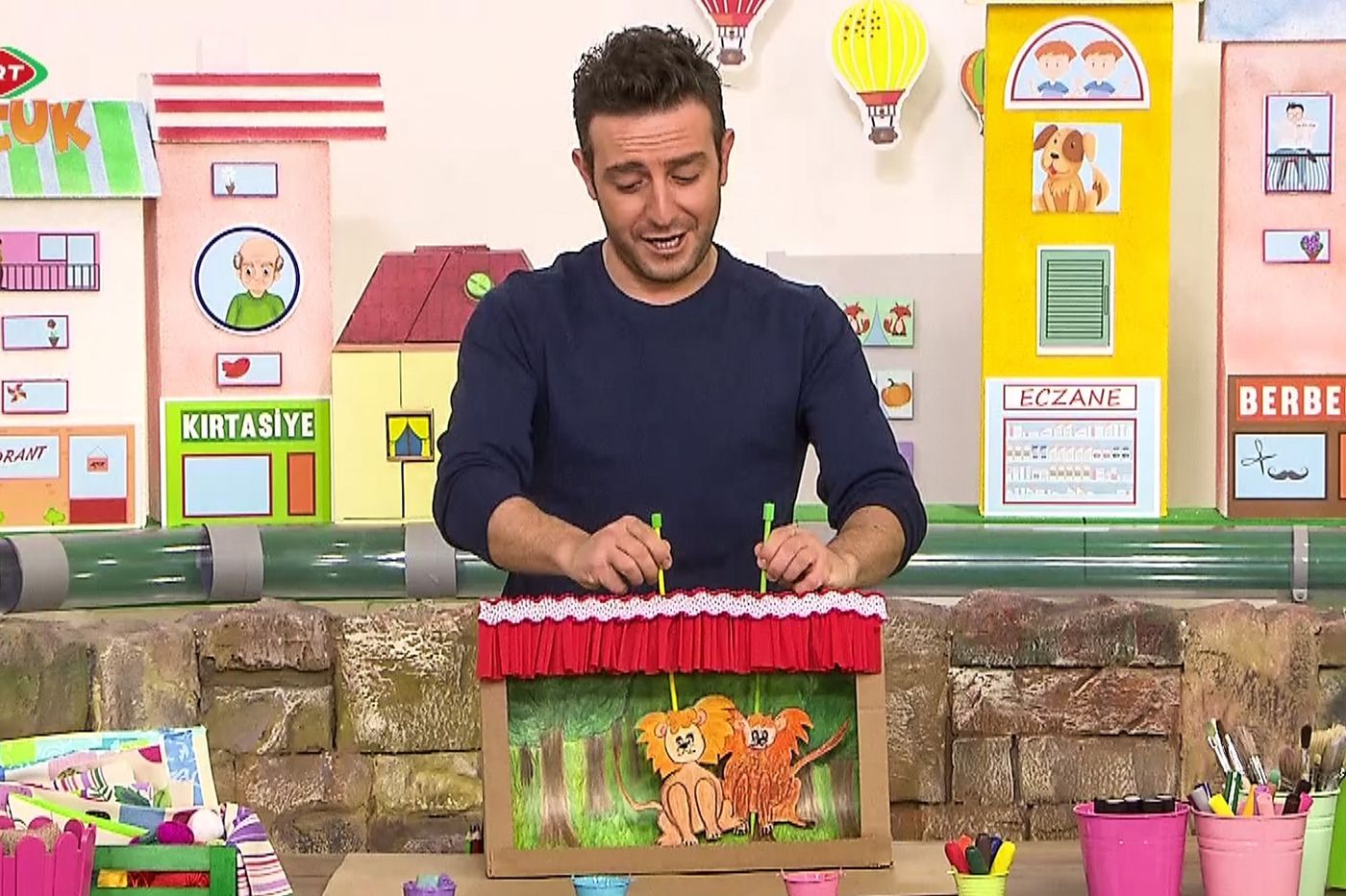 Programdan kare: sunucu masadaki kırmızı perdeli karton kukla sahnesinde sopalı kuklaları oynatıyor
