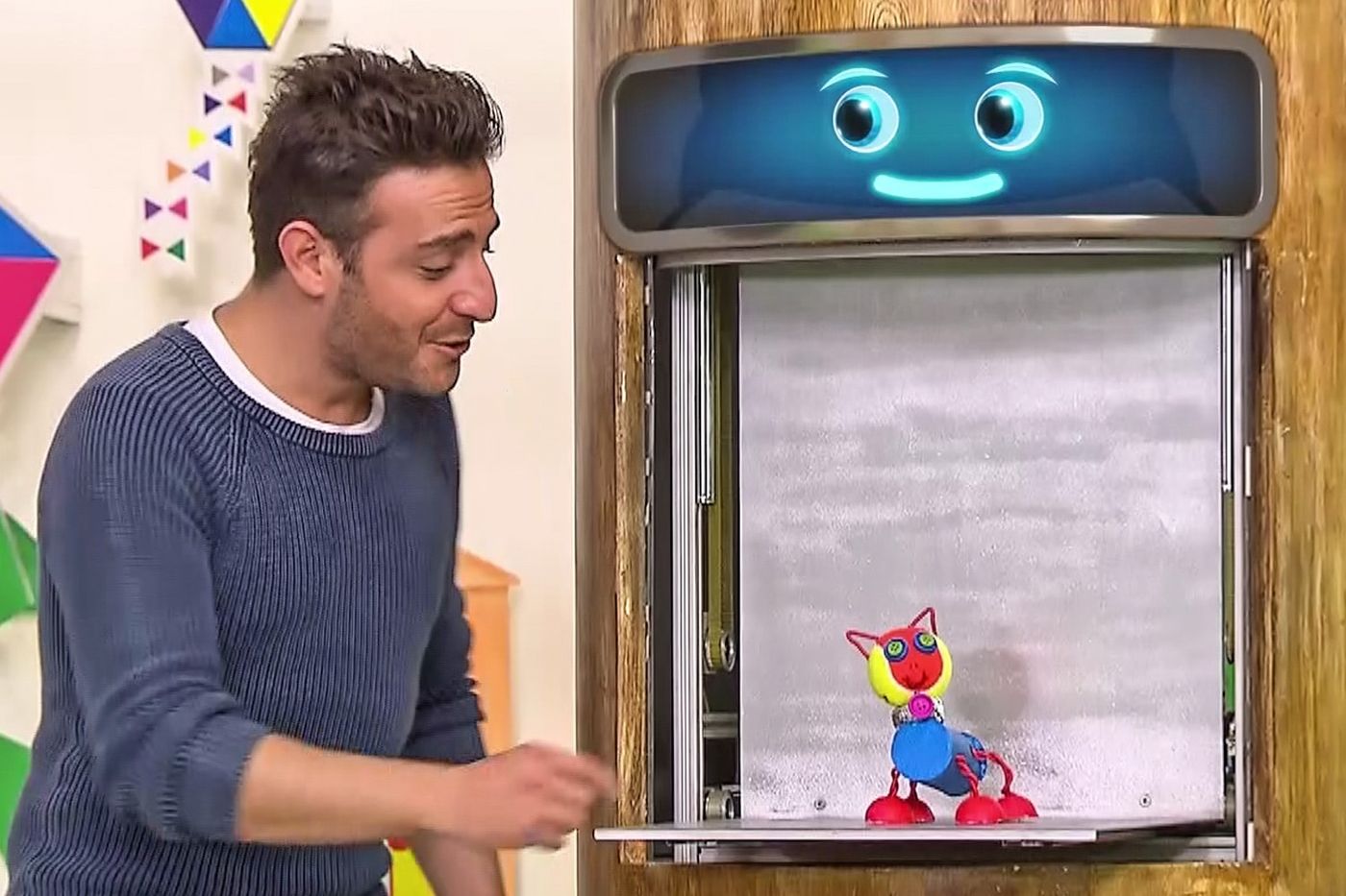 Yayından kare: sunucu stüdyo duvarındaki ekranlı dolapta duran renkli rulo robota bakıyor