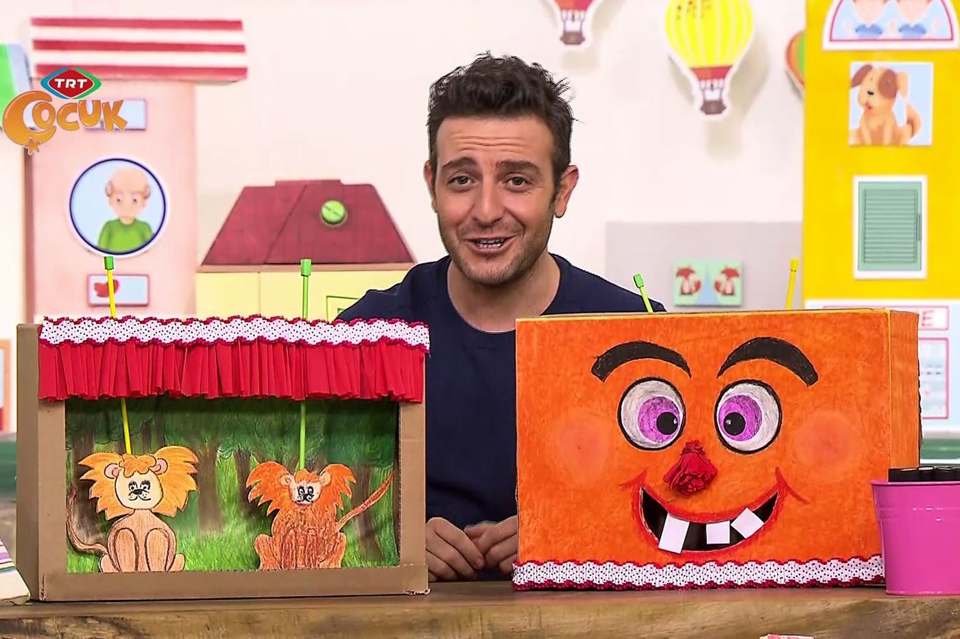 Kukla sahnesi yapılan programdan kare: sunucu karton kutudan ağaçlı sahne ve turuncu yüzlü kutu ile