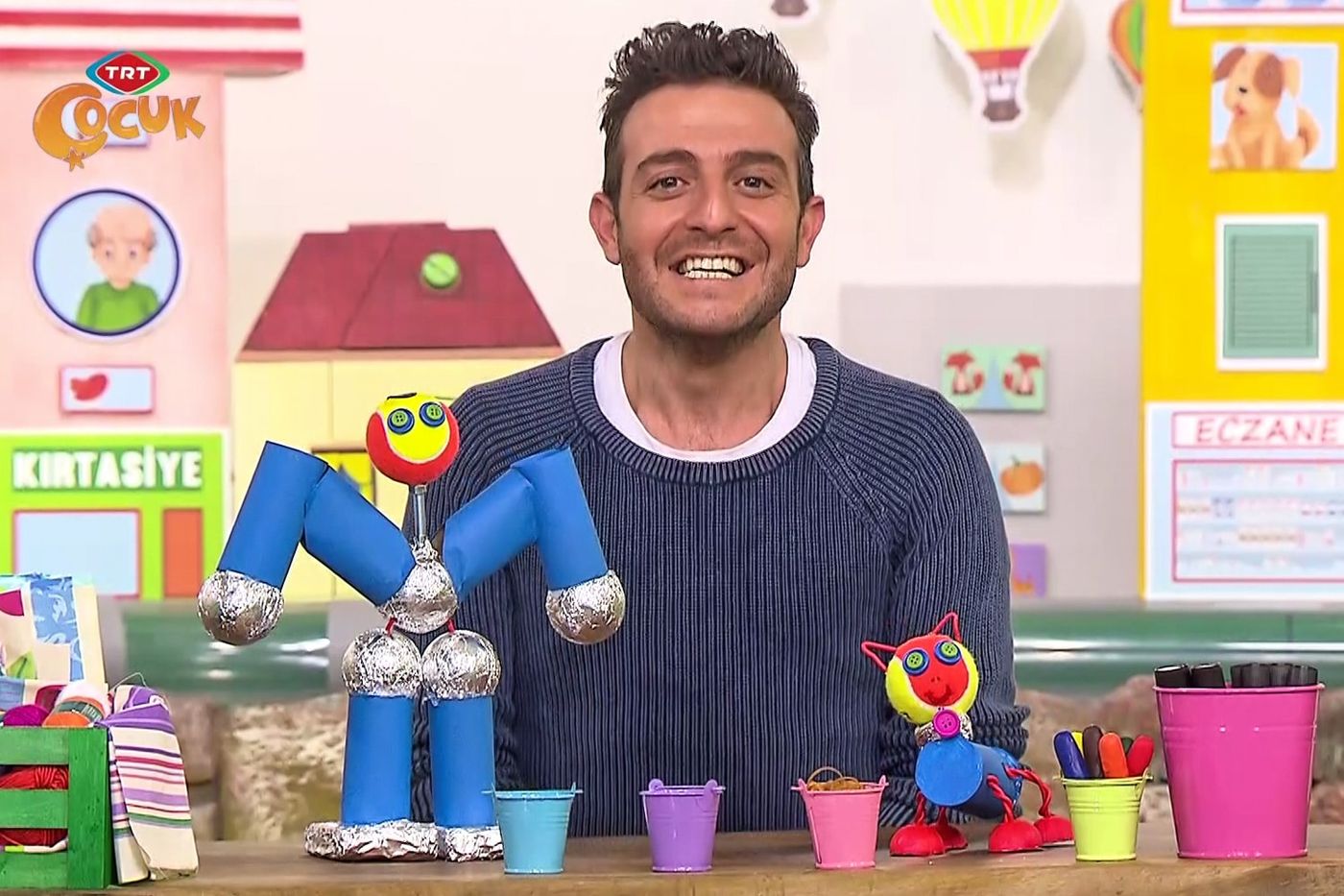 Rulo robot yayın karesi: sunucu masada mavi rulolardan ve folyo eklemlerden robotun yanında