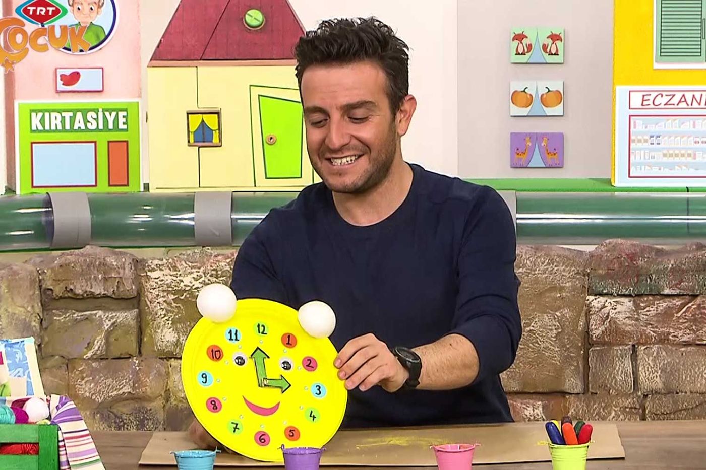 Televizyon programından kare: sunucu stüdyo masasında sarı tabaktan yapılmış gülen yüzlü saati gösteriyor