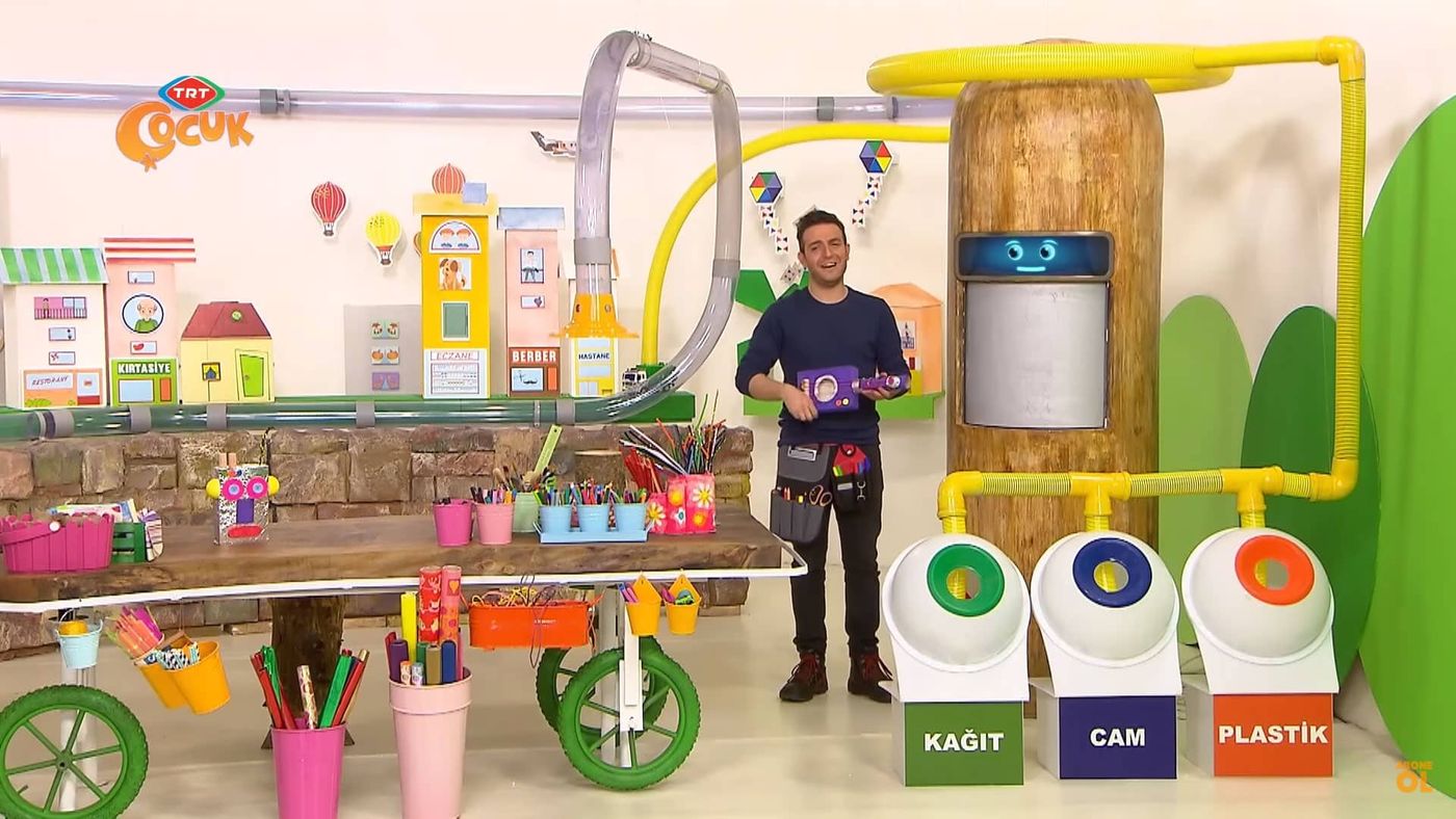Yayından geniş plan: stüdyoda sunucu, önünde malzeme masası ve kağıt, cam, plastik geri dönüşüm kutuları