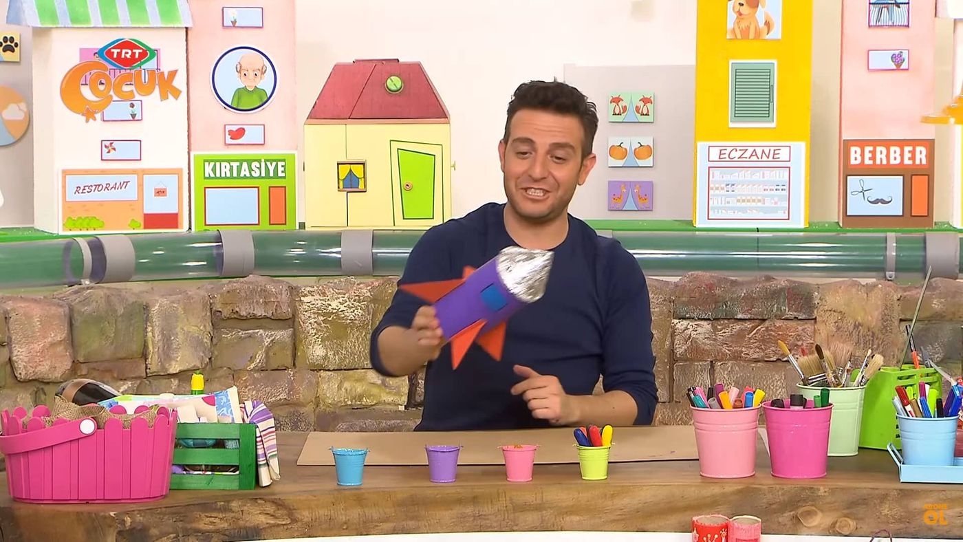 Video karesi: sunucu renkli atölye masasının başında bitmiş füze kalemliği gösteriyor