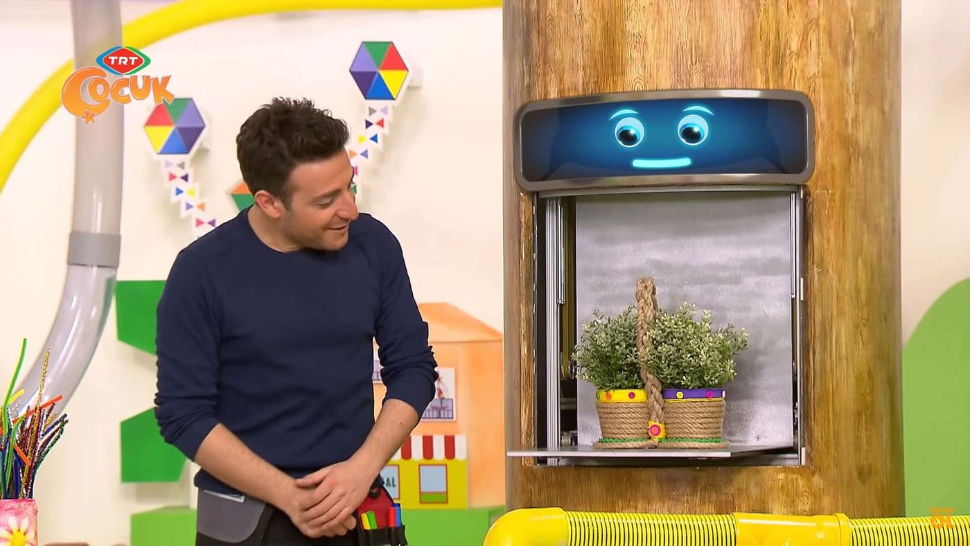 Televizyon programından kare: sunucu stüdyoda, yanındaki pencerede ip sarılı ikili saksı duruyor