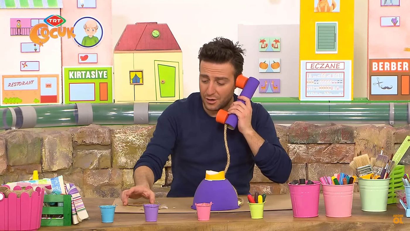 Programdan kare: sunucu kağıt rulo ahizeyi kulağına götürüyor, önünde mor oyuncak telefon