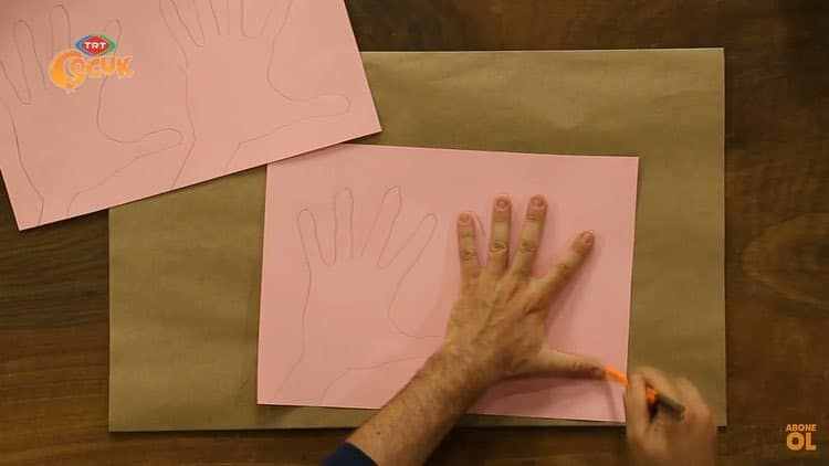 Kraft kağıt üstündeki pembe kağıda el konup çevresi kalemle çiziliyor