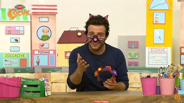 Televizyon programından kare: sunucu kedi kulaklı taç ve çizili bıyıklarla masada anlatıyor
