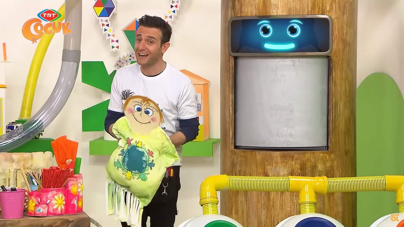 Sunucu, boyalı tişört gövdeli ve karton yüzlü bebeği kucağında tutarak gösteriyor