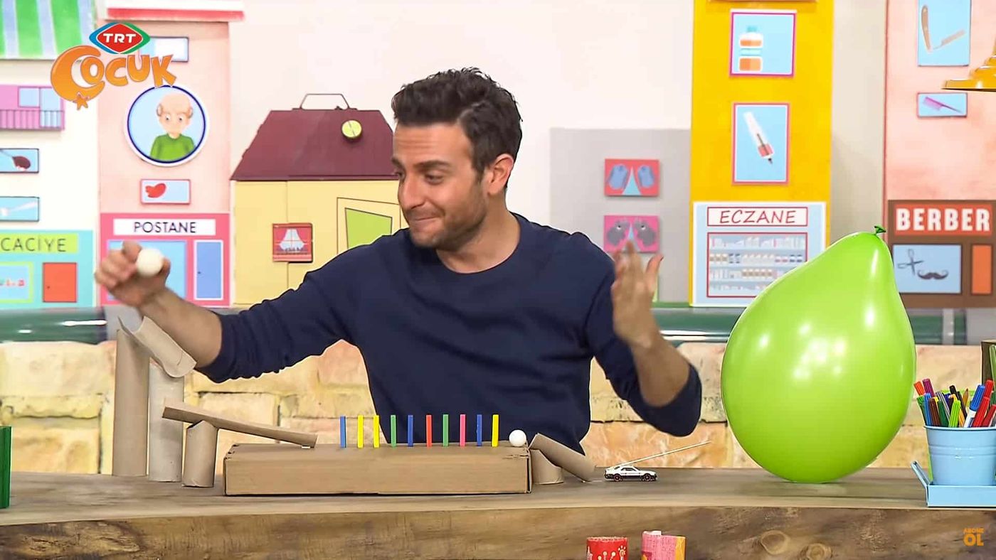 Programdan kare: sunucu masadaki zincirleme düzeneğin başında, yanında şişirilmiş yeşil balon