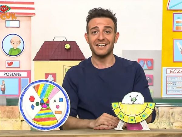 Televizyon programından kare: sunucu desenli kağıt tabak frizbi ve sarı kağıt tabaktan uzay aracı gösteriyor