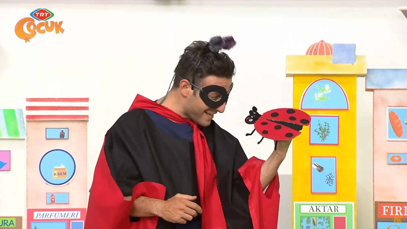 Programdan kare: maskeli ve pelerinli uğur böceği kostümüyle sunucu, elinde kağıt uğur böceği