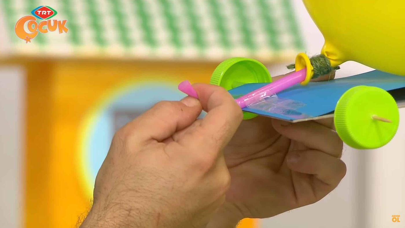 Pembe pipete bağlanmış sarı balon, mavi karton gövdeli ve yeşil şişe kapağı tekerlekli arabaya tutturuluyor