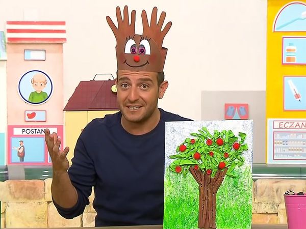 Geyik taç video karesi: sunucu başında el izi boynuzlu karton taç, yanında yeşil yapraklı ağaç tablosu