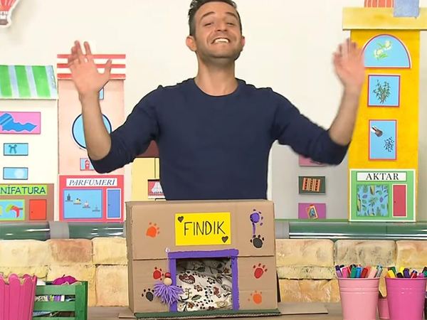 Kedi Evi televizyon programından kare: el sallayan sunucu, önünde pati izli karton kutu kedi evi