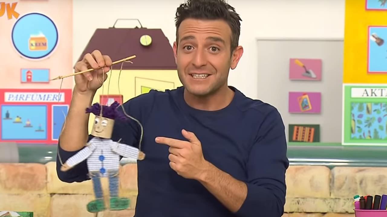 Televizyon programından kare: sunucu elindeki ipli karton kuklayı parmağıyla işaret ediyor