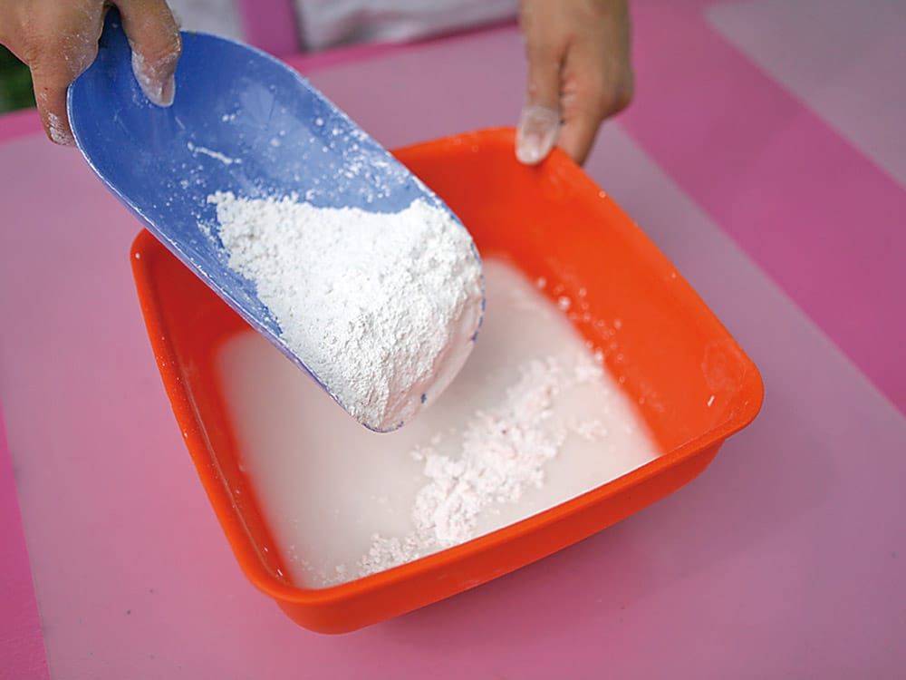 Bir el kepçeyle beyaz tozu turuncu plastik kaba döküyor, oyun tahtası hazırlığının ilk adımı