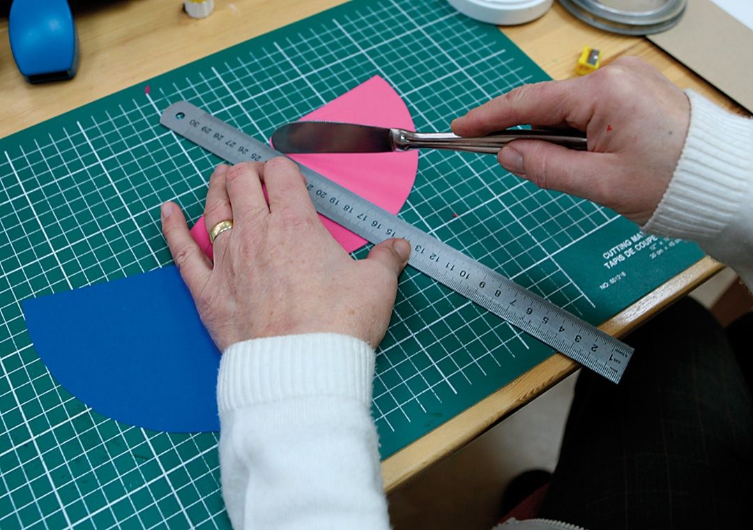 Cetvel ve maket bıçağıyla pembe yarım daire kartonun ortasına katlama çizgisi çizen eller